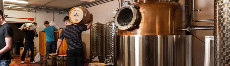 People working in distillery, with barrels and metal equipment.